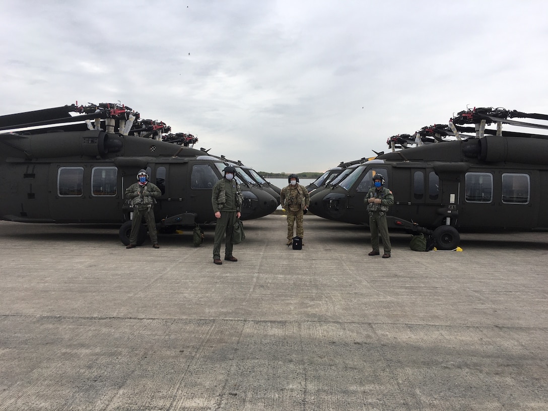 Four military officers in flight suits stand in front of U.S. Army UH-60 Blackhawk helicopters.