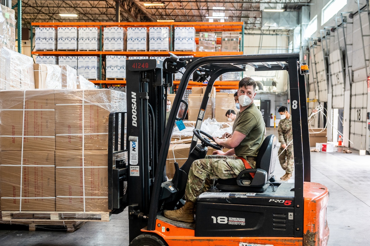 A soldier drives a forklift lifting a pallet of boxes.
