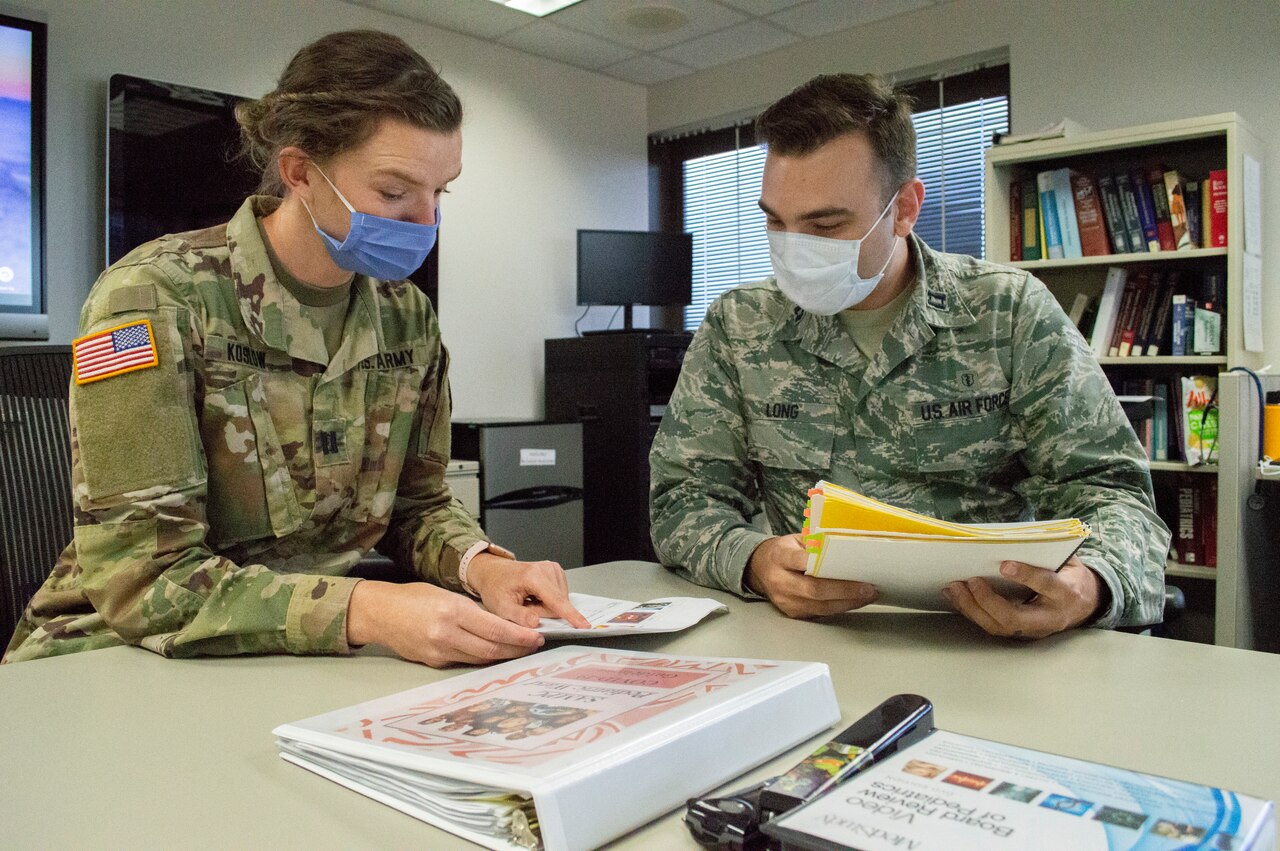 Two military doctors wearing face masks sit at a table comparing notes.
