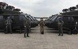 Four soldiers in flight suits stand in front of UH-60 Blackhawk helicopters.