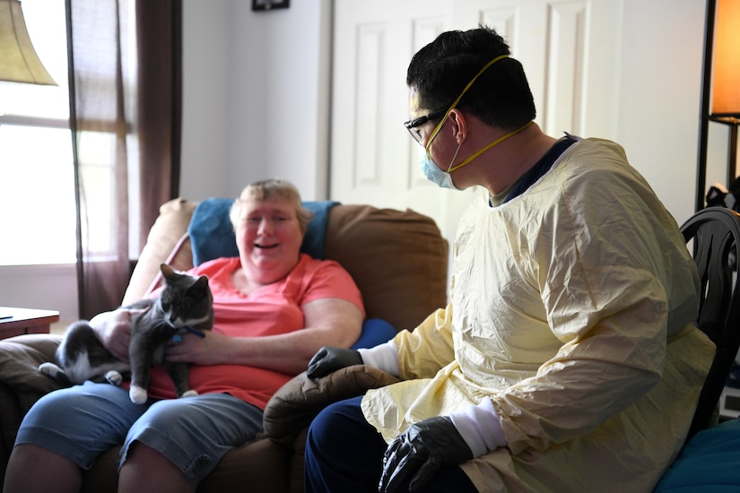 An airman wearing personal protective equipment sits and chats with a woman who is seated and holding a cat on her lap.