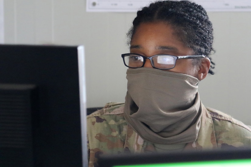 A soldier wearing a face mask and eyeglasses works at a computer.
