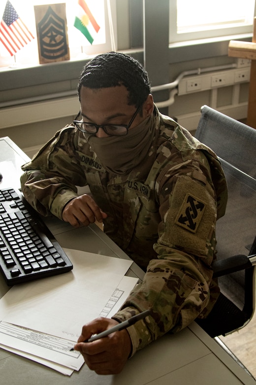 A soldier wearing a face mask and eyeglasses is seated at a table, taking notes as he works on an online assignment.