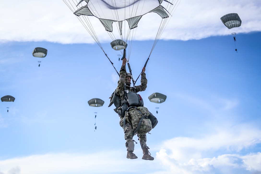 A soldier holds onto his parachute while descending in the blue sky, as other parachutes descend in the background.