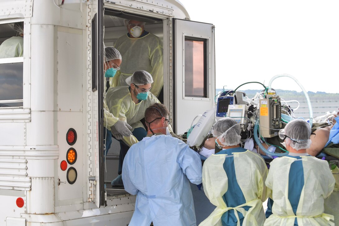 Airmen in protective gear move a patient from the back of an open bus to a flightline.