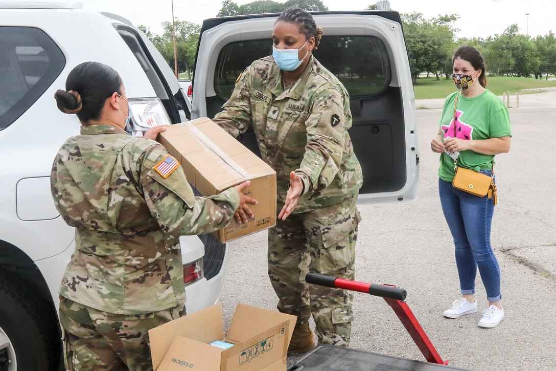 A soldier hands a cardboard box to another soldier standing by the open trunk of a vehicle as a civilian looks on.