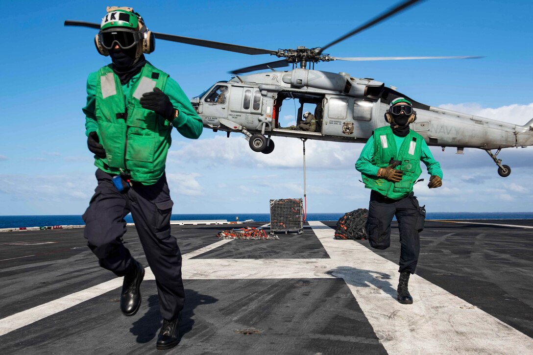 Two sailors run away from a helicopter that is lifting off from the deck of a ship.