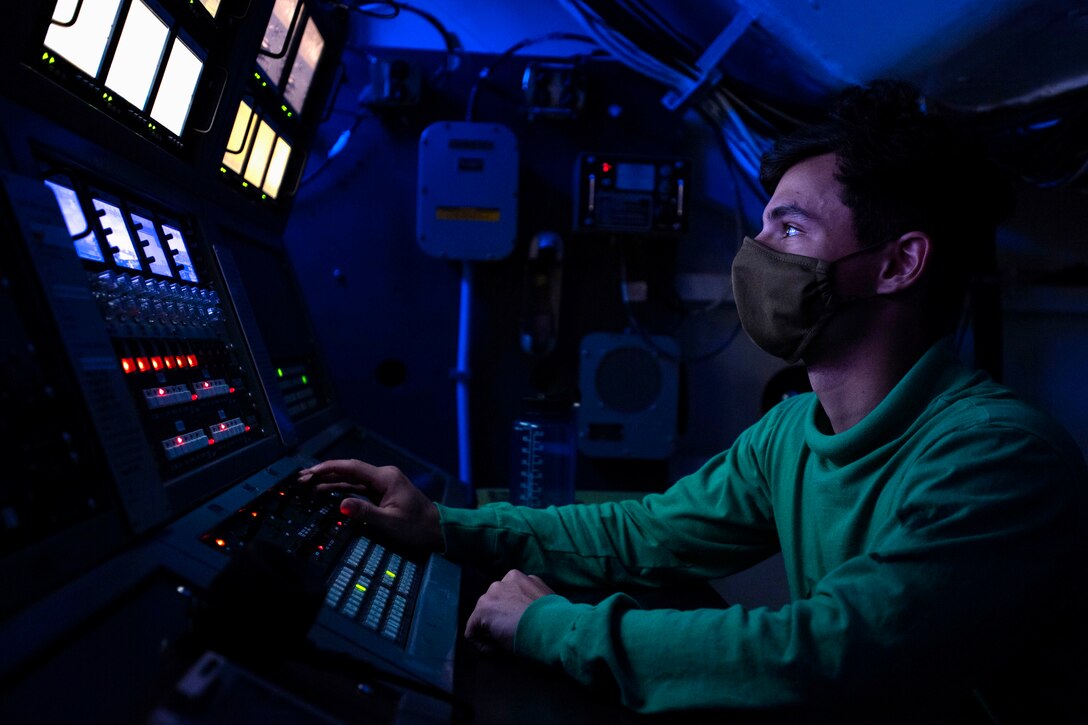 A sailor illuminated by blue light sits in front of monitors.