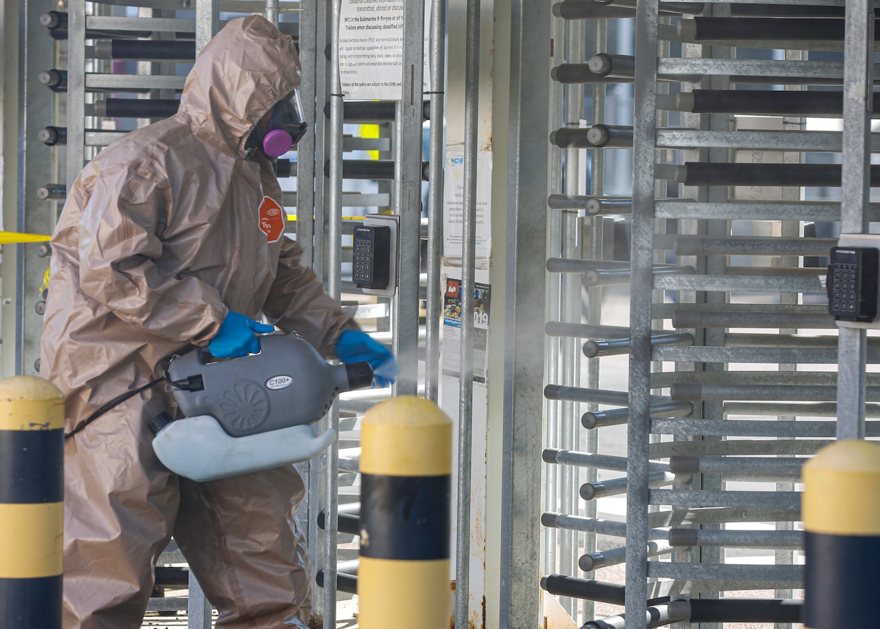 A man dressed in full protective gear sprays a turnstile entrance with disinfectant.