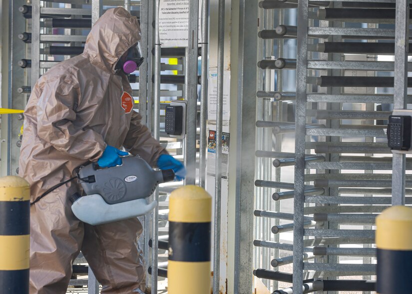 A man dressed in full protective gear sprays a turnstile entrance with disinfectant.
