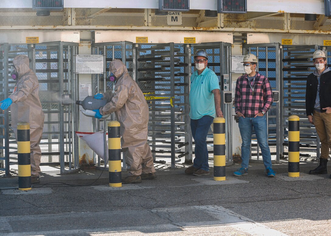 Two men in full protective equipment sanitize turnstiles as three other men wearing face masks stand nearby.