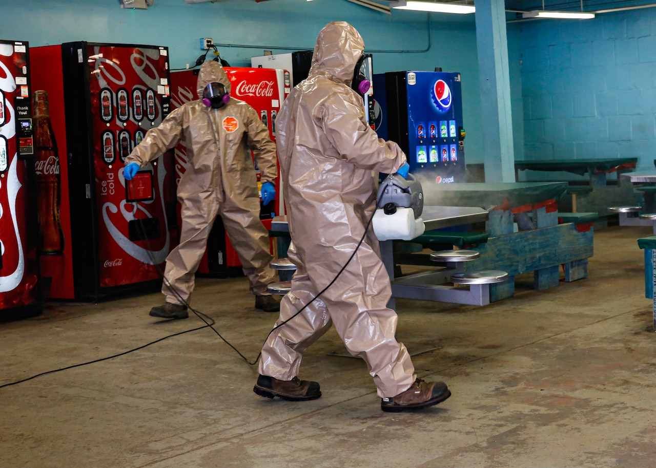 Men dressed in full protective gear spray a room with disinfectant.