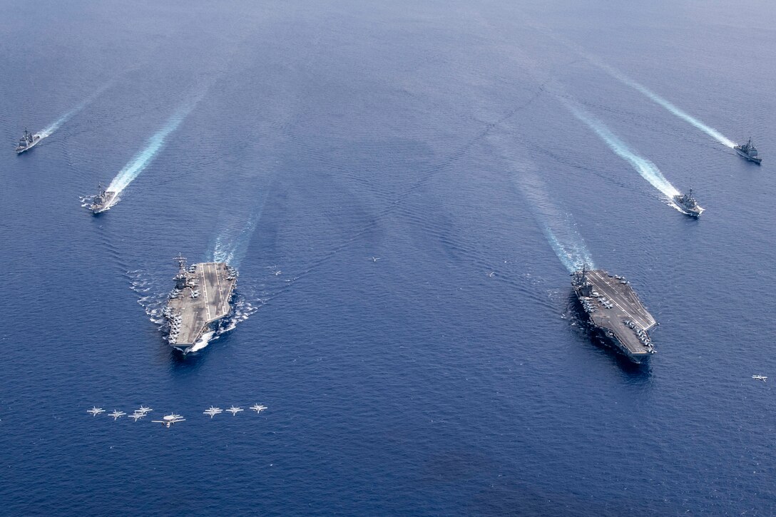 Military aircraft fly in formation over a group of military ships in the ocean.