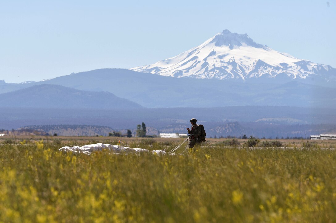 An airman stands in a field with a parachute with a mountain in the background.