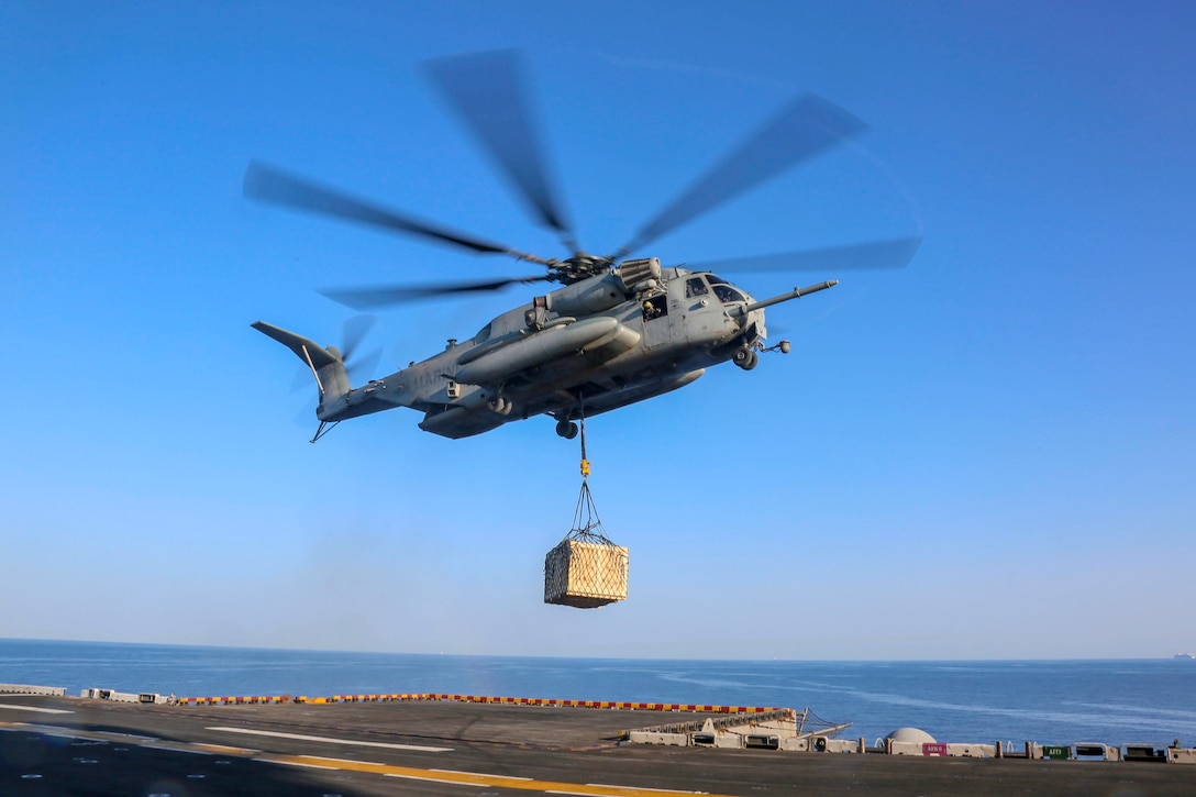 A helicopter carries cargo above the deck of a ship.