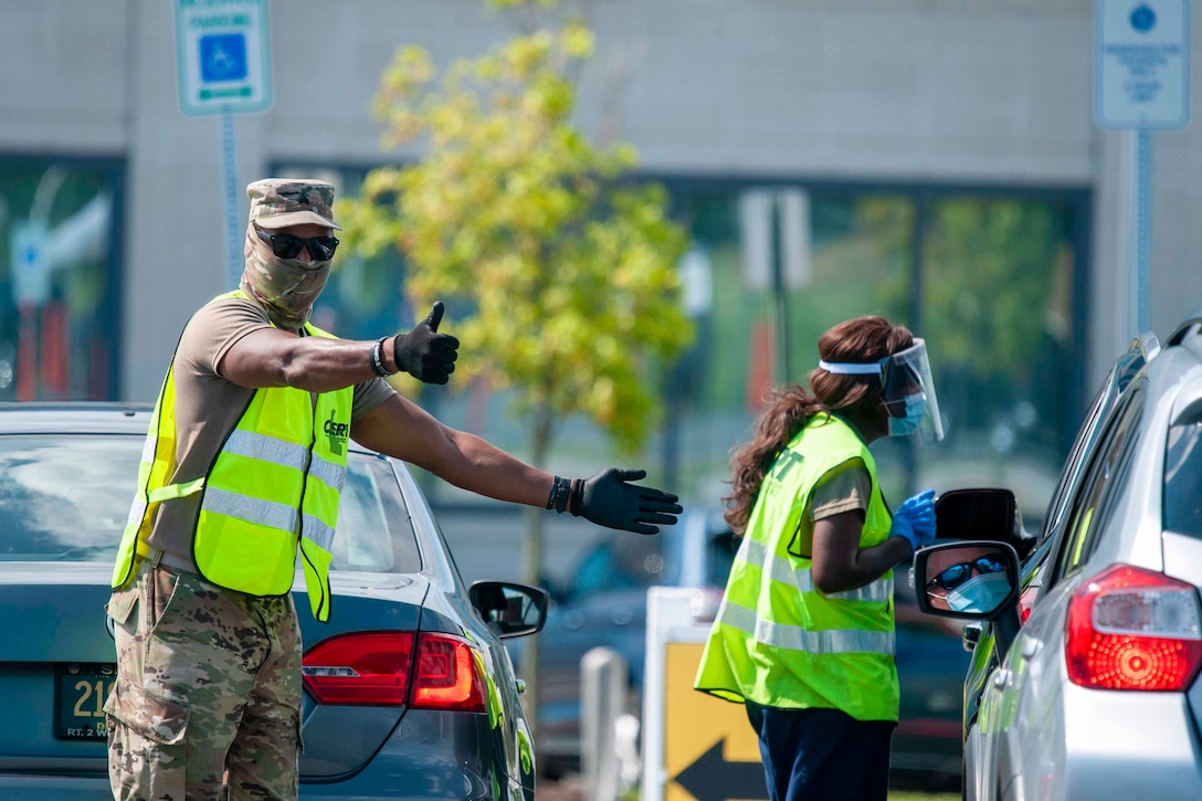 A soldier wearing personal protective equipment signals toward cars.
