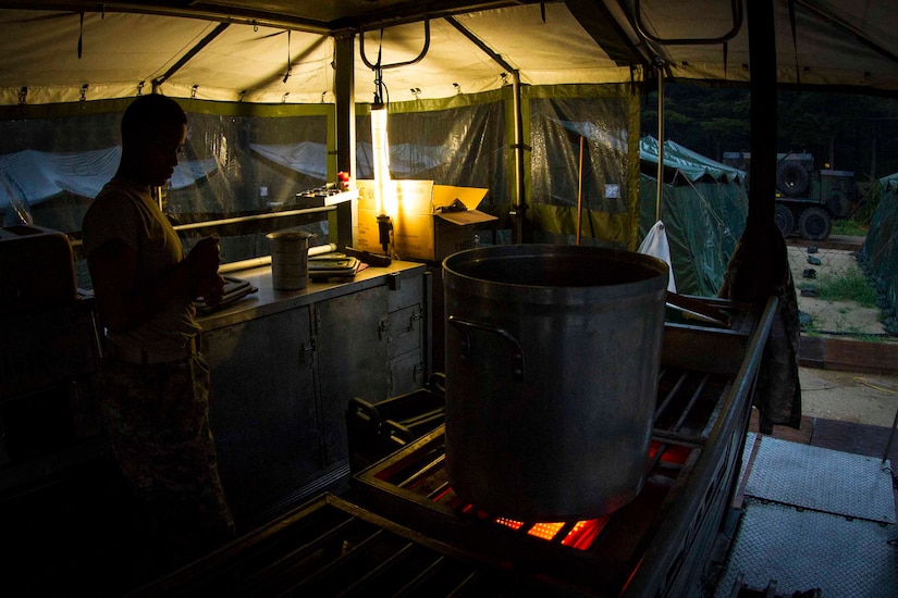 A soldier watches a large, metal container heat over an outdoor stove.