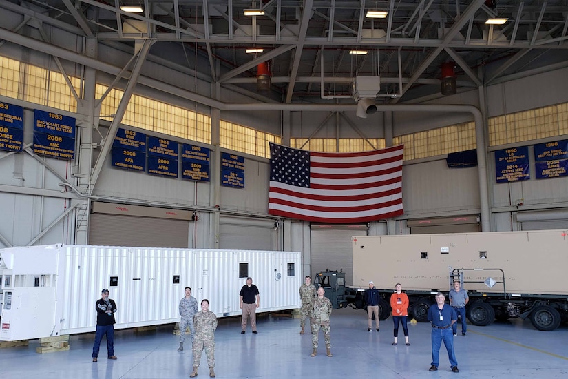 Airmen and civilians stand in a hangar beneath a large U.S. flag.