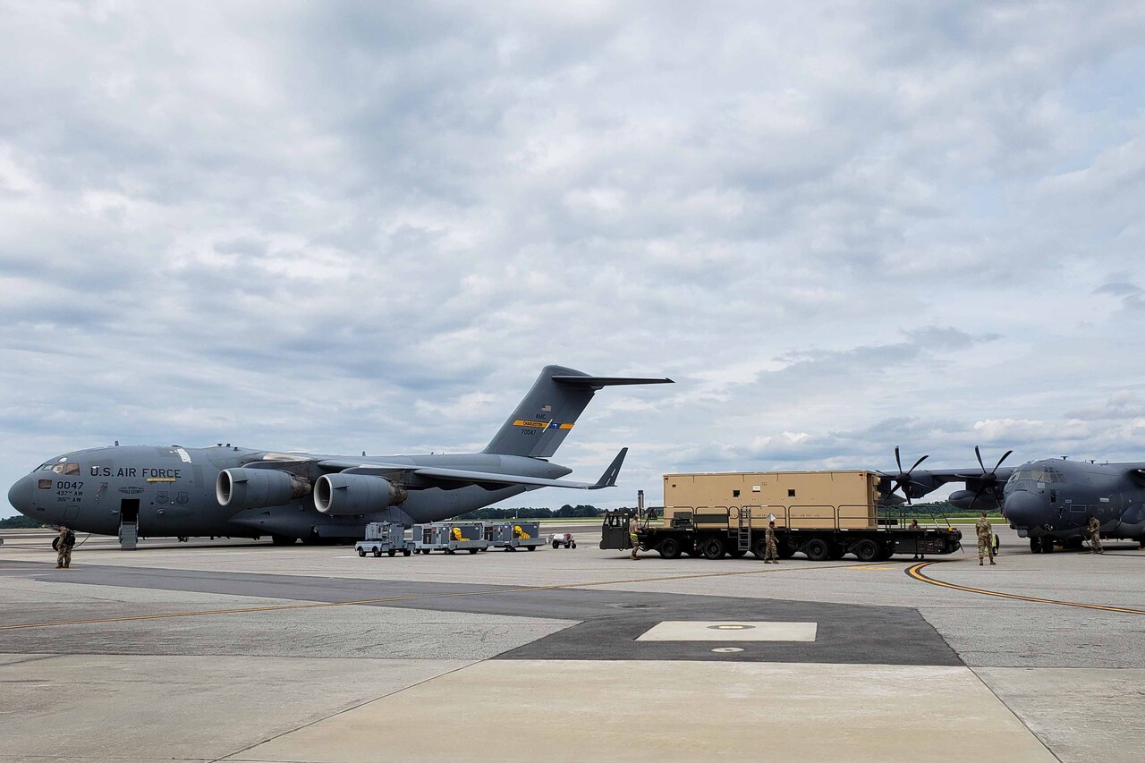 Two transport aircraft on a ramp with a large, multiwheeled flatbed vehicle between them.