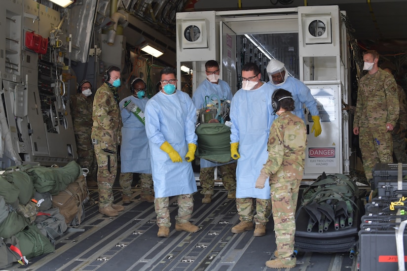 Medical personnel, some in military uniforms, gather outside a large metal box that is situated inside a large military aircraft.