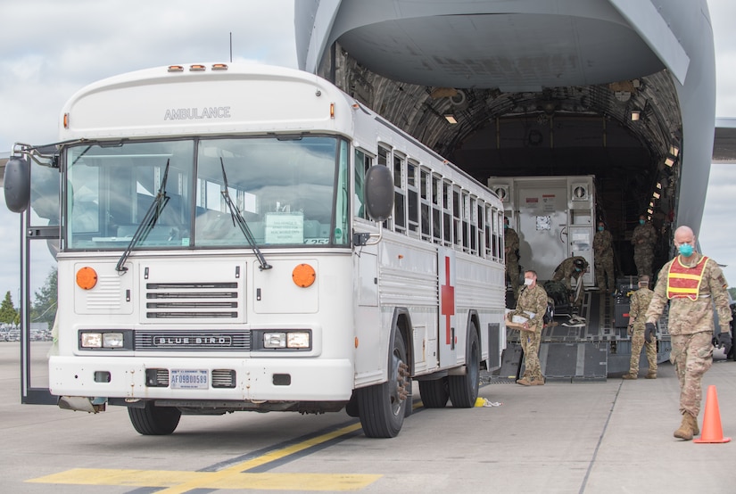 A white bus with a red cross on its side backs up to a large military aircraft.