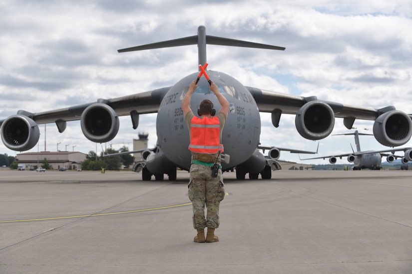 A service member stands on a flight line and uses orange-colored wands to direct the movement of a large military aircraft.