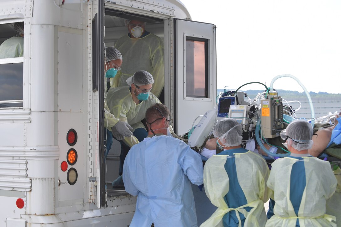 Medical personnel move a patient on a stretcher out of the back of a white bus.