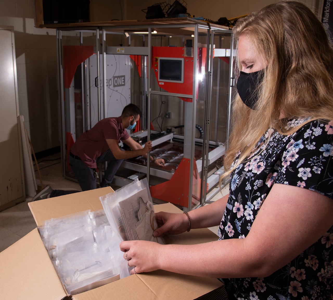 A woman wearing a mask places personal protective equipment into a plastic bag as a man in the background works at a 3D printer.