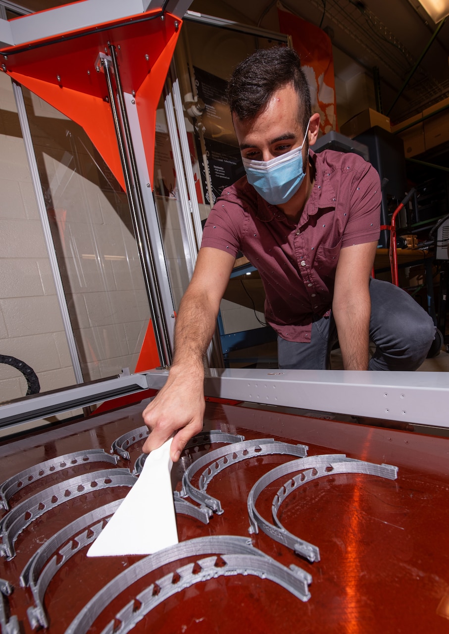 A man wearing a mask uses a spatula-type tool to collect 3D-printed personal protective equipment parts.