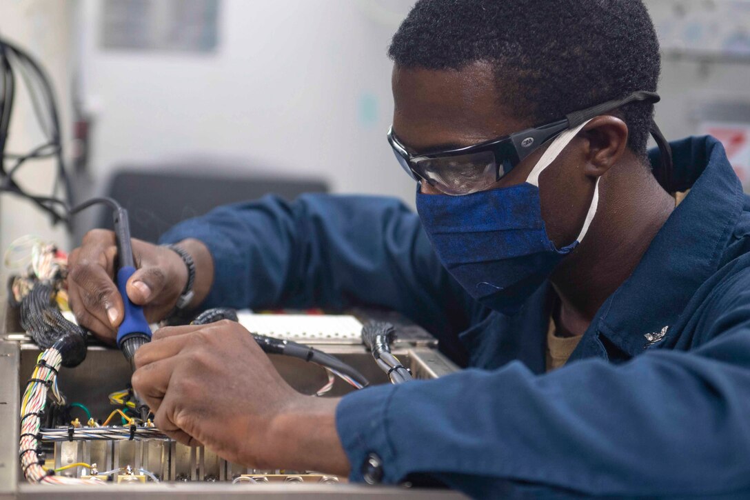 A sailor wearing a face mask solders a piece of equipment.