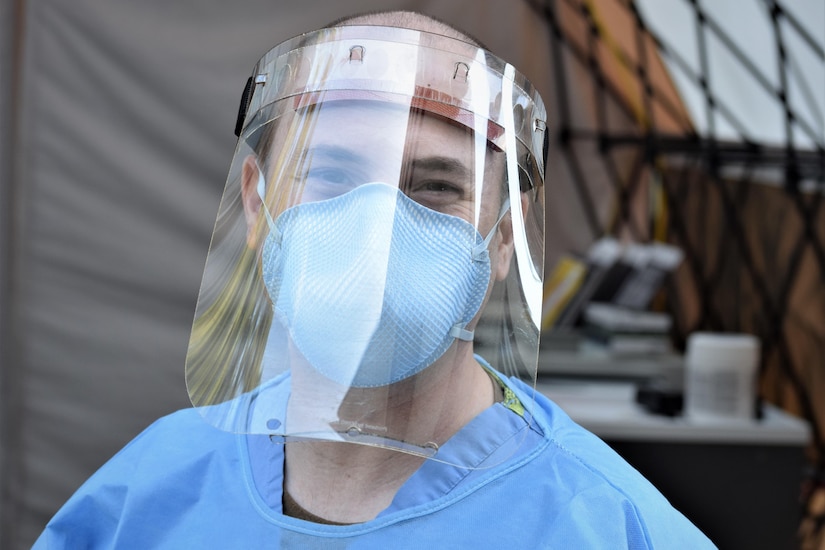 A sailor poses for a head-and-shoulders shot while wearing surgical scrubs, a face mask and a face shield.