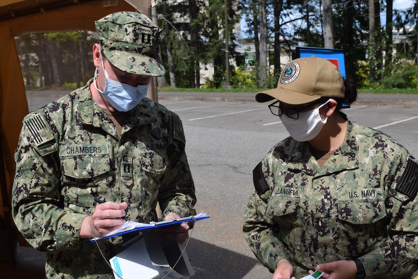 Two sailors wearing face masks and camouflage uniforms compare notes.