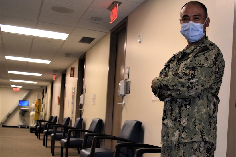 A sailor wearing a face mask and a camouflage uniform stands with his arms folded in a medical waiting room.
