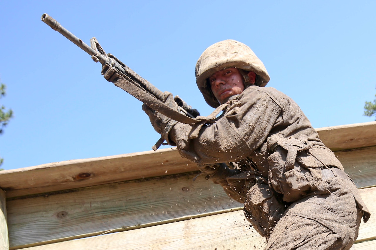 A Marine with a rifle. Part of an obstacle course is in the background.