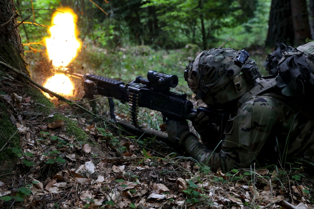 A soldier in camouflage fires a weapon while lying on the ground.