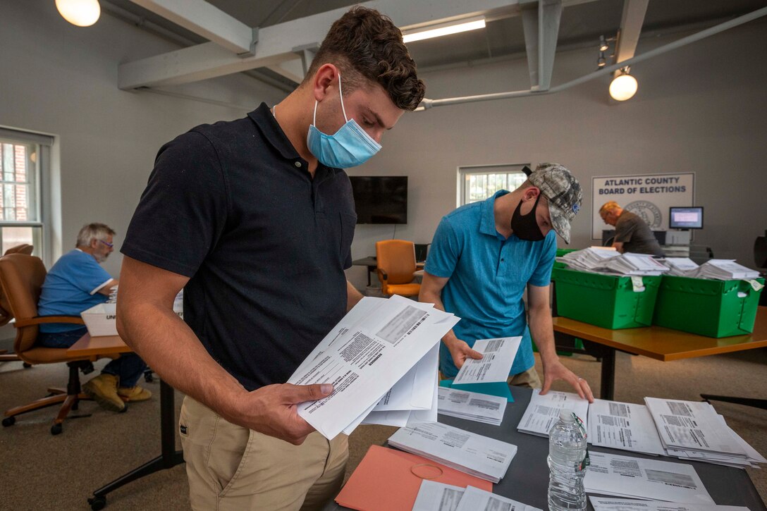 A soldier wearing a mask and another soldier behind him help alphabetize ballots.