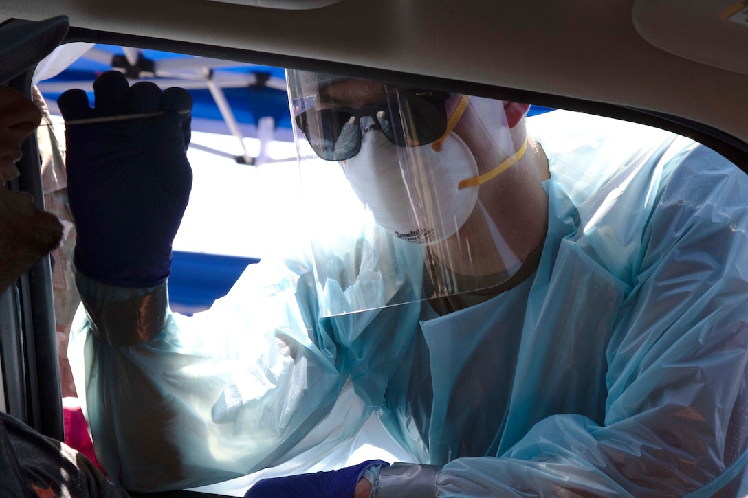 A guardsman wearing personal protective equipment administers a COVID-19 test on a passenger through a car window.