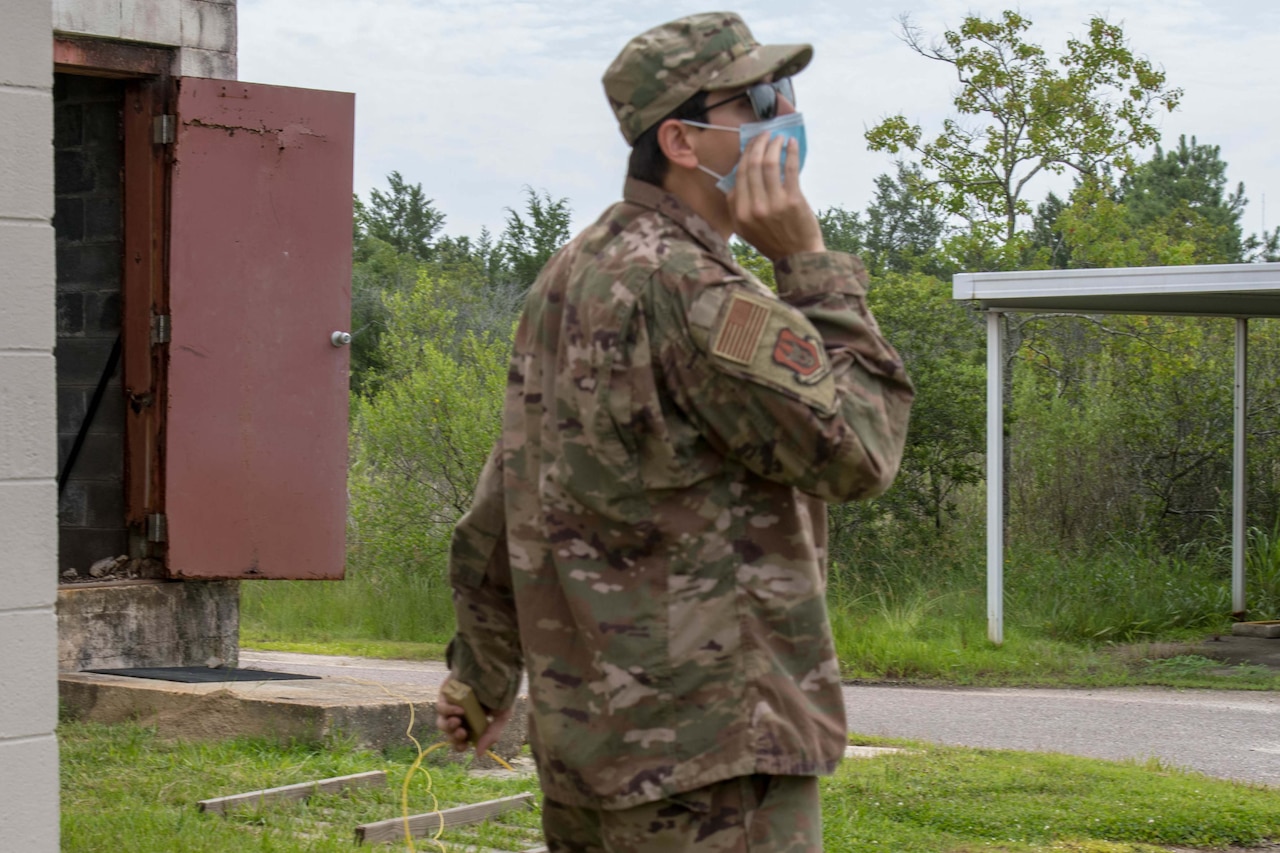 An airman wearing a face mask and holding some yellow wire in his left hand cups his right hand to his face to yell an instruction.