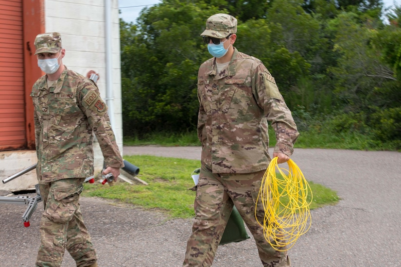 Airmen wearing face masks walk side by side. One is carrying equipment, and the other is carrying a rolled-up length of yellow wire.