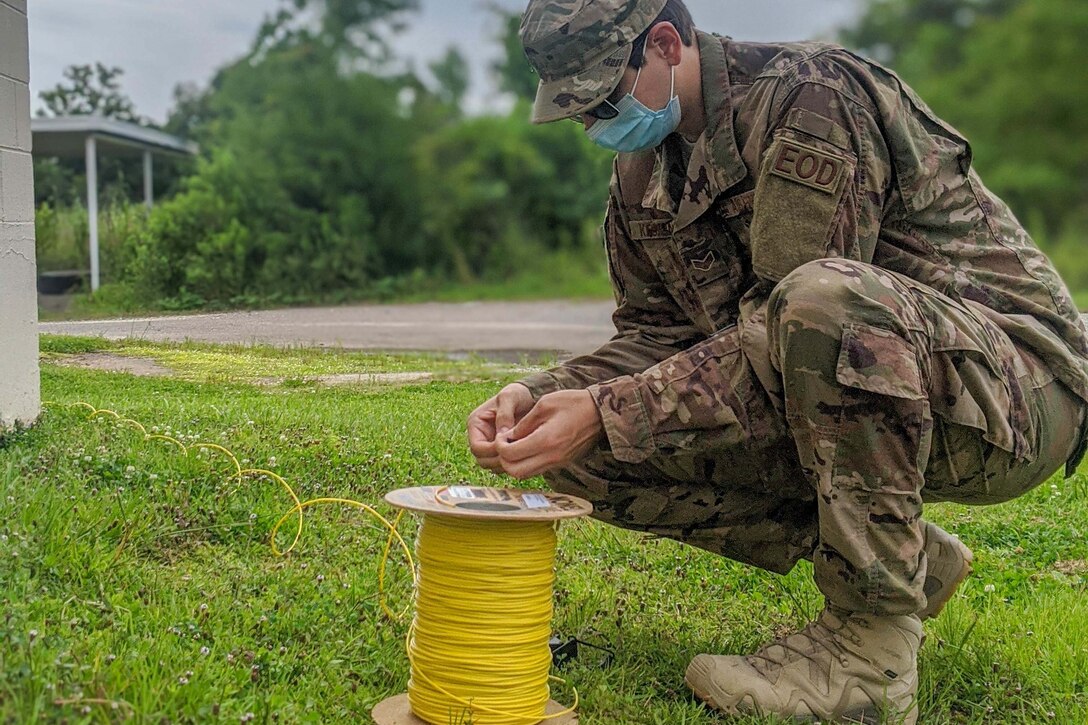 An airman wearing a face mask kneels next to a large spool of yellow wire.
