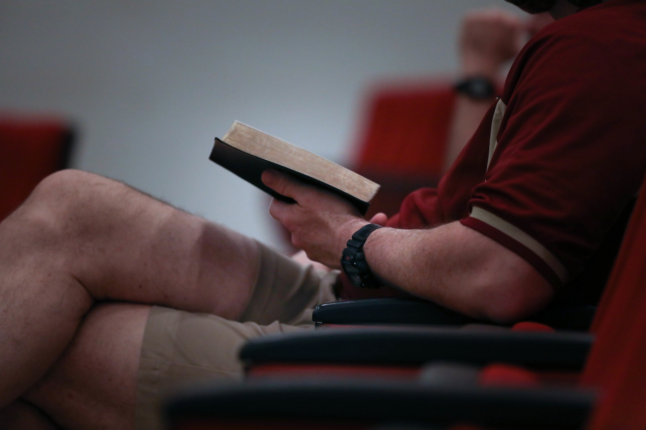 A man seated in a pew holds a book open during a chapel service.