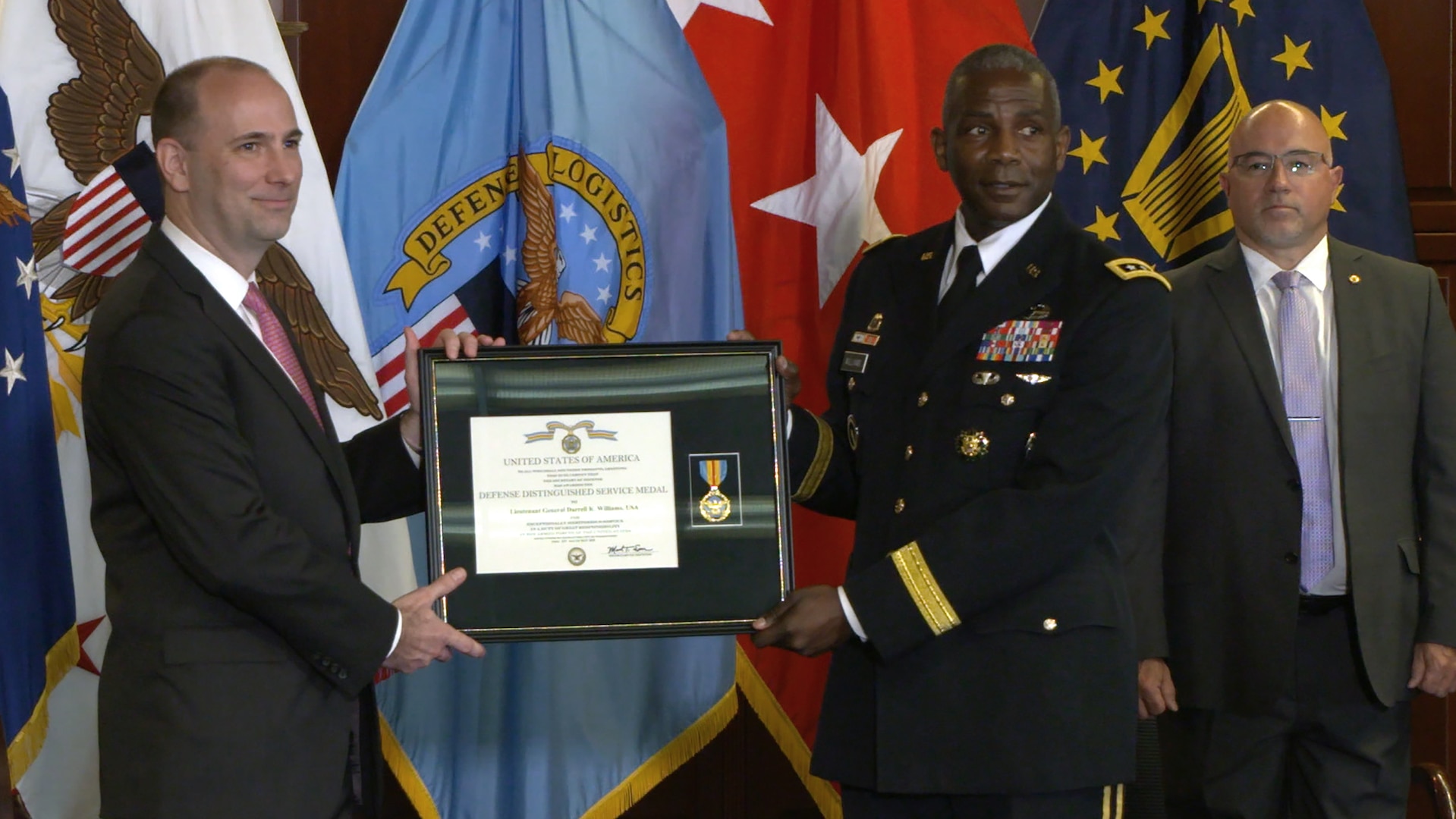 White man in suit and black man in Army dress uniform hold a framed certificate and medal in front of U.S. and DLA flags