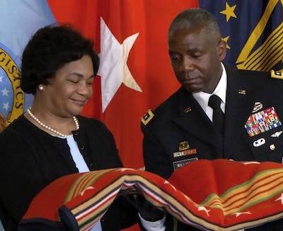Black man and woman stand in front of the U.S., DLA and 3-star general flags holding a red blanket with multicolored stars and stripes.