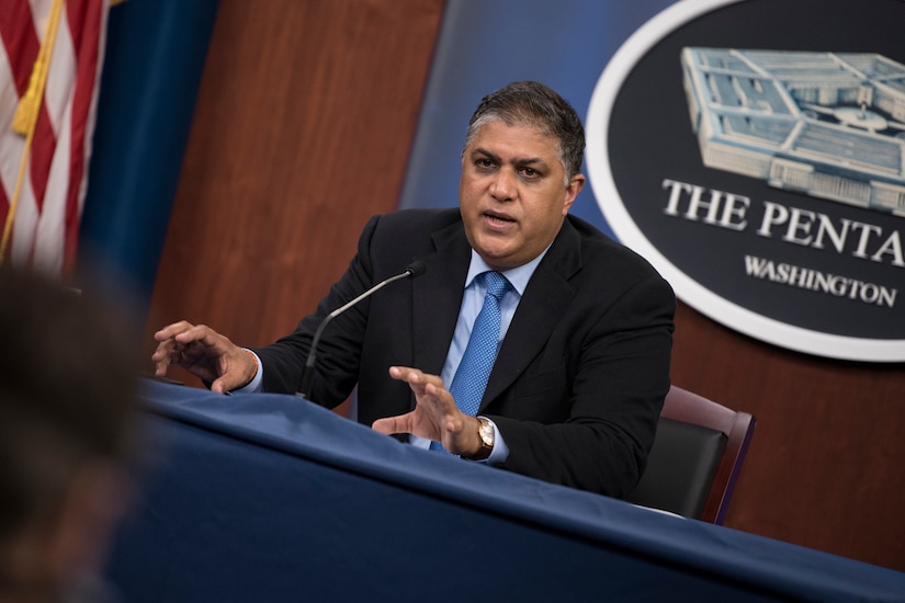 A man sits in front of a microphone. Behind him is a sign on the wall that says The Pentagon.