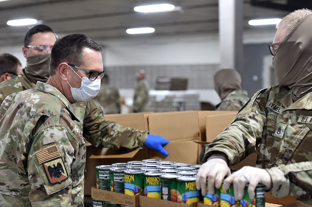 Several service members wearing face masks check the contents of a case  of canned goods.