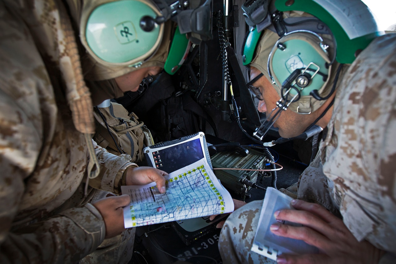 Two service members wearing aviation headgear, crouch near each other and look at a map.