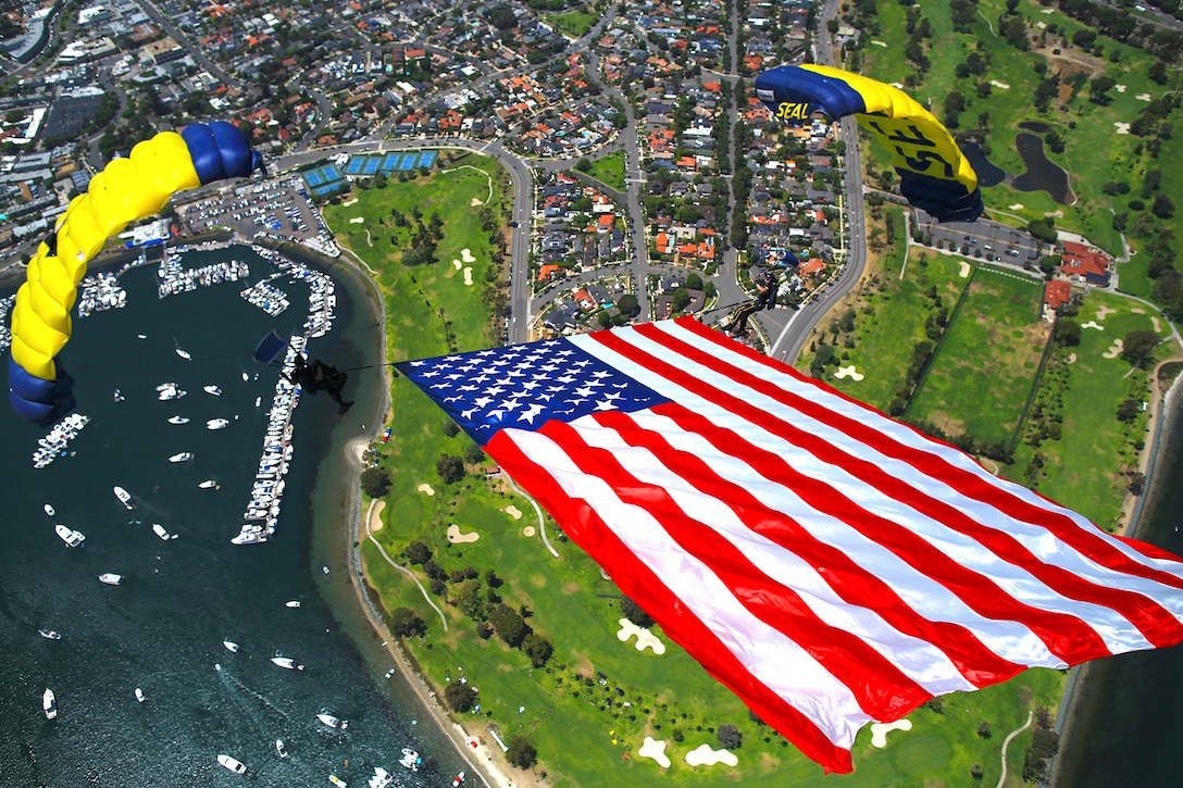 Sailors free fall while holding up the American flag over a city.