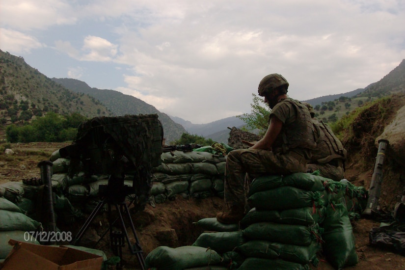 Two soldiers sit on sandbags. Mountains are in the background.