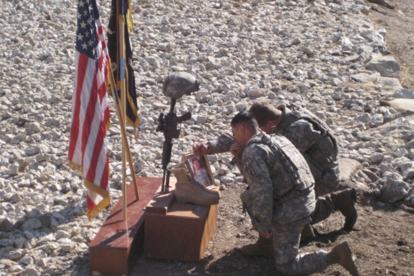 Two soldiers kneel in front of a memorial for a fallen comrade.