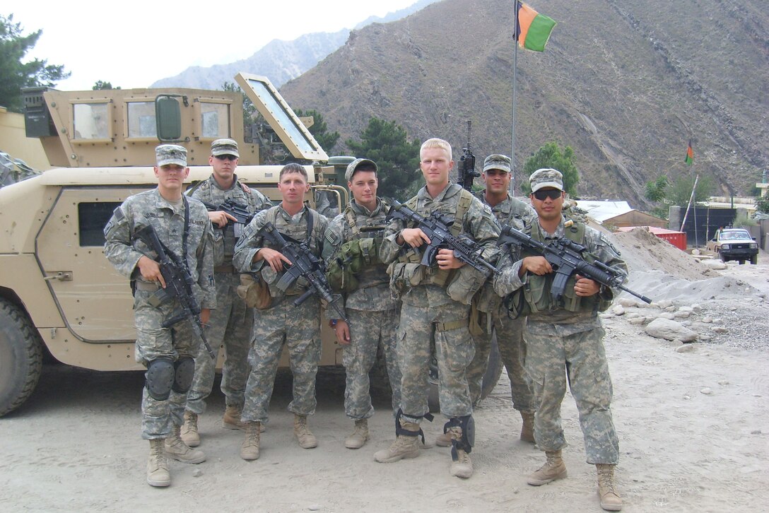 Seven soldiers pose near a tank beside a mountain.